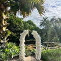 White floral archway in a garden setting with palm trees and a clear sky.