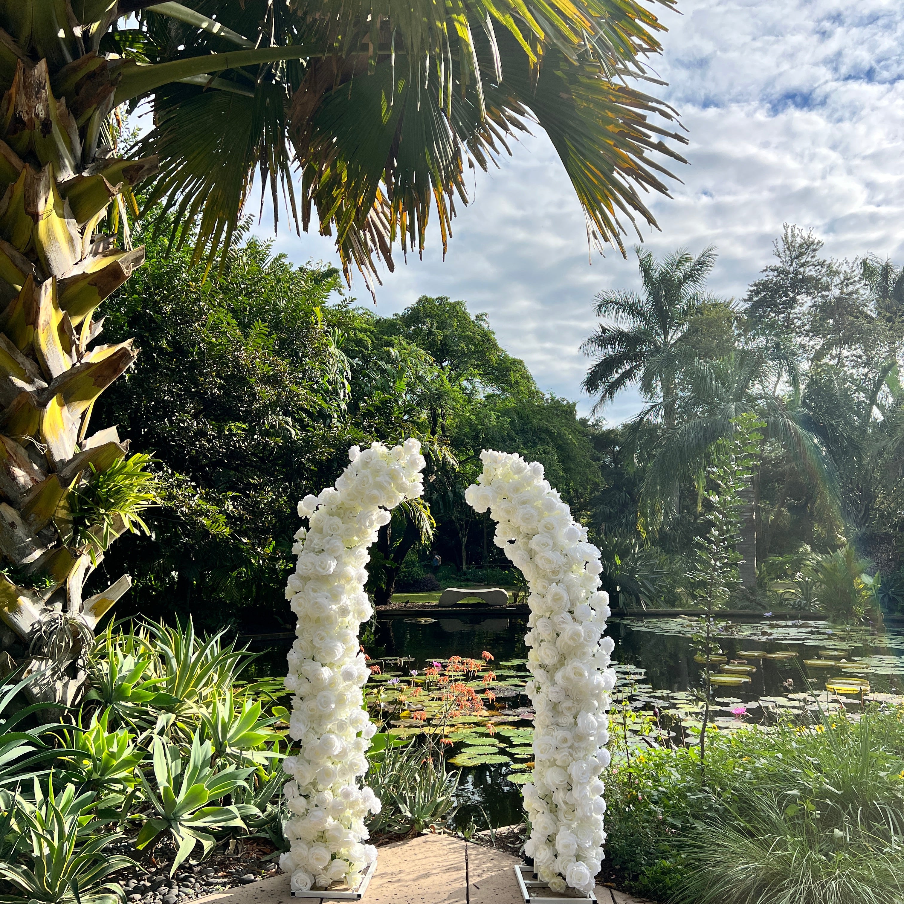 White floral archway in a garden setting with palm trees and a clear sky.