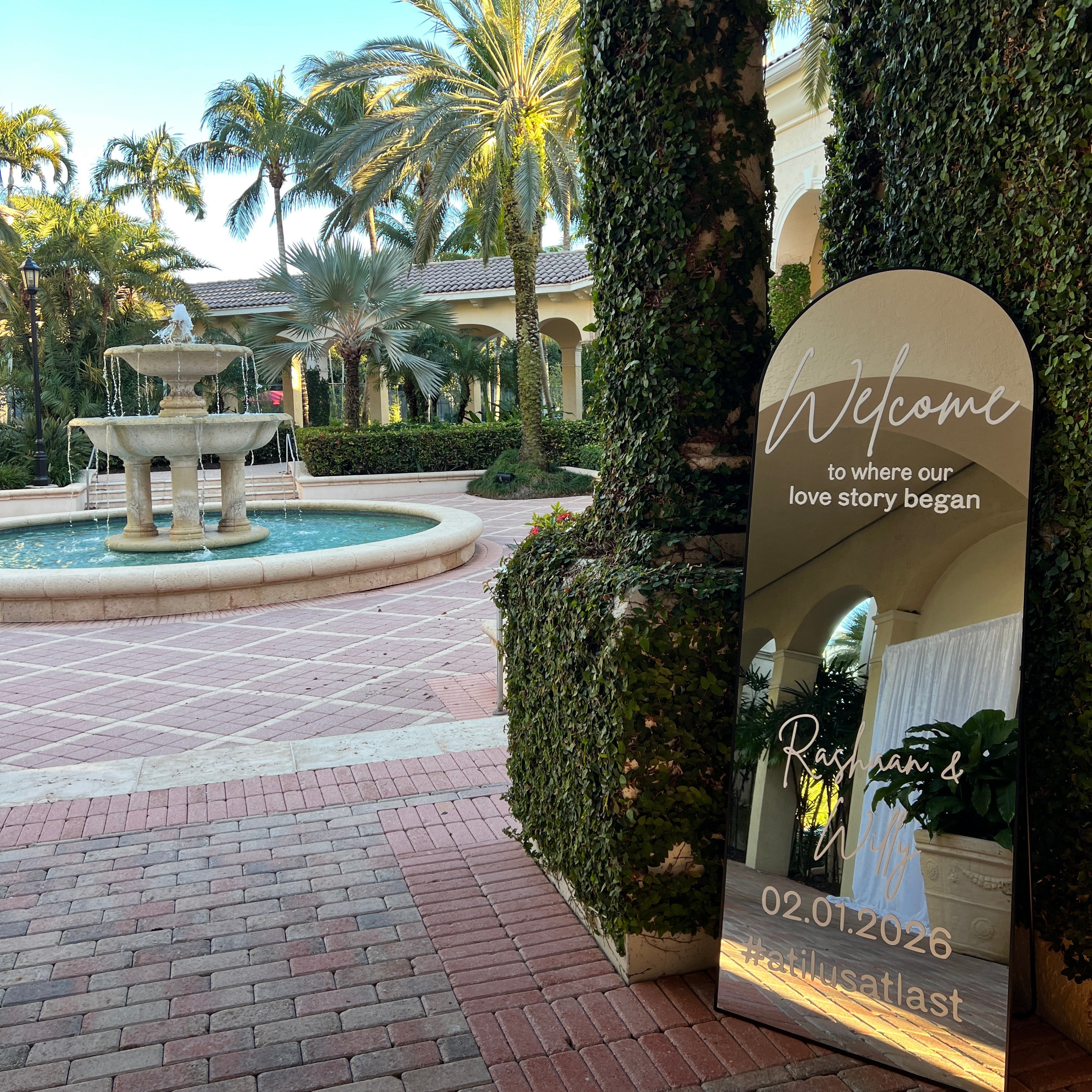 Mirror with text and contact information in a garden setting with palm trees and a fountain.