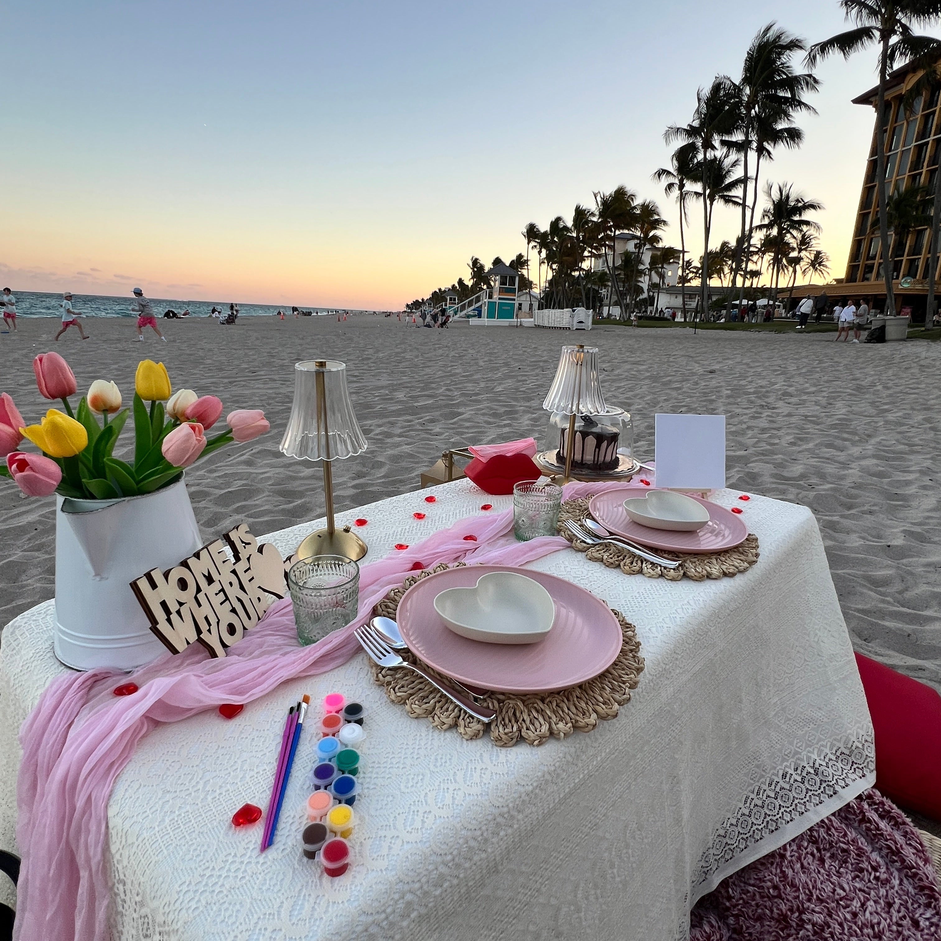 Dining table setup with pink tablecloth, plates, and flowers on a beach at sunset.