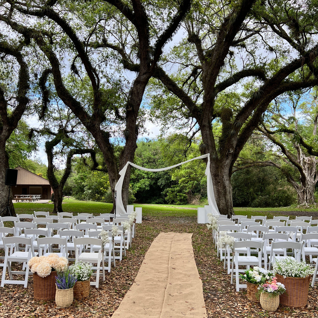 Outdoor wedding setup with white chairs, floral arrangements, and a tree in the background.