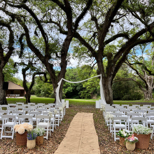 Outdoor wedding setup with white chairs, floral arrangements, and a tree in the background.